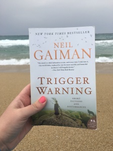 Hand holding Trigger Warning by Neil Gaiman in front of a beach with the ocean in the background